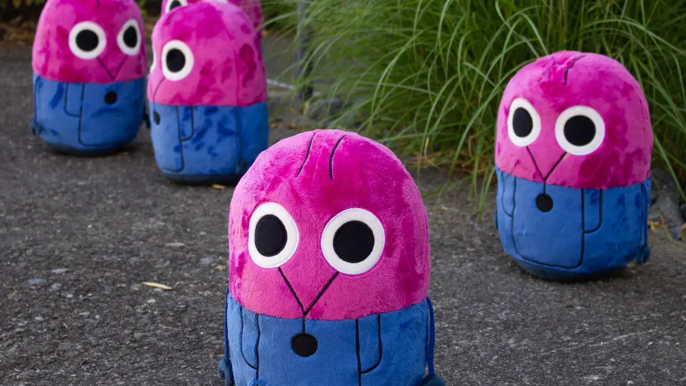 Fünf pink-blaue iosys-Maskottchen als Plüschfiguren auf Asphalt vor Gräsern